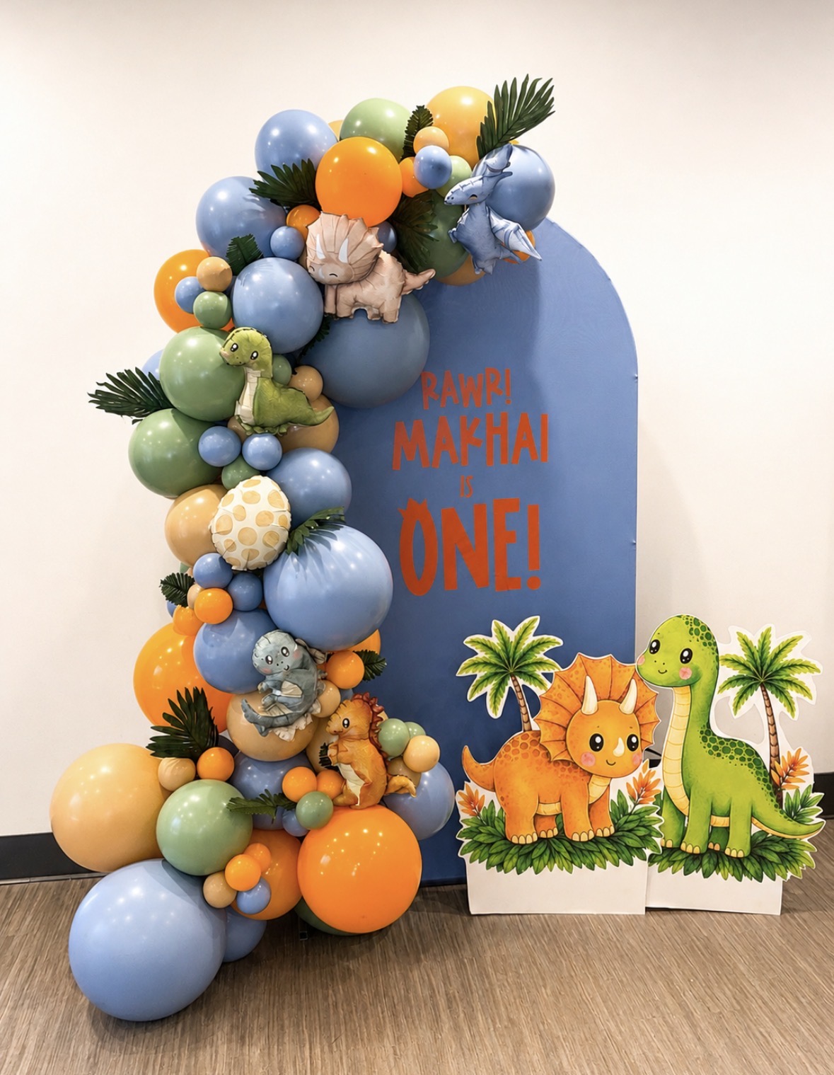 Blue, green, and orange dinosaur first birthday backdrop with balloon garland
